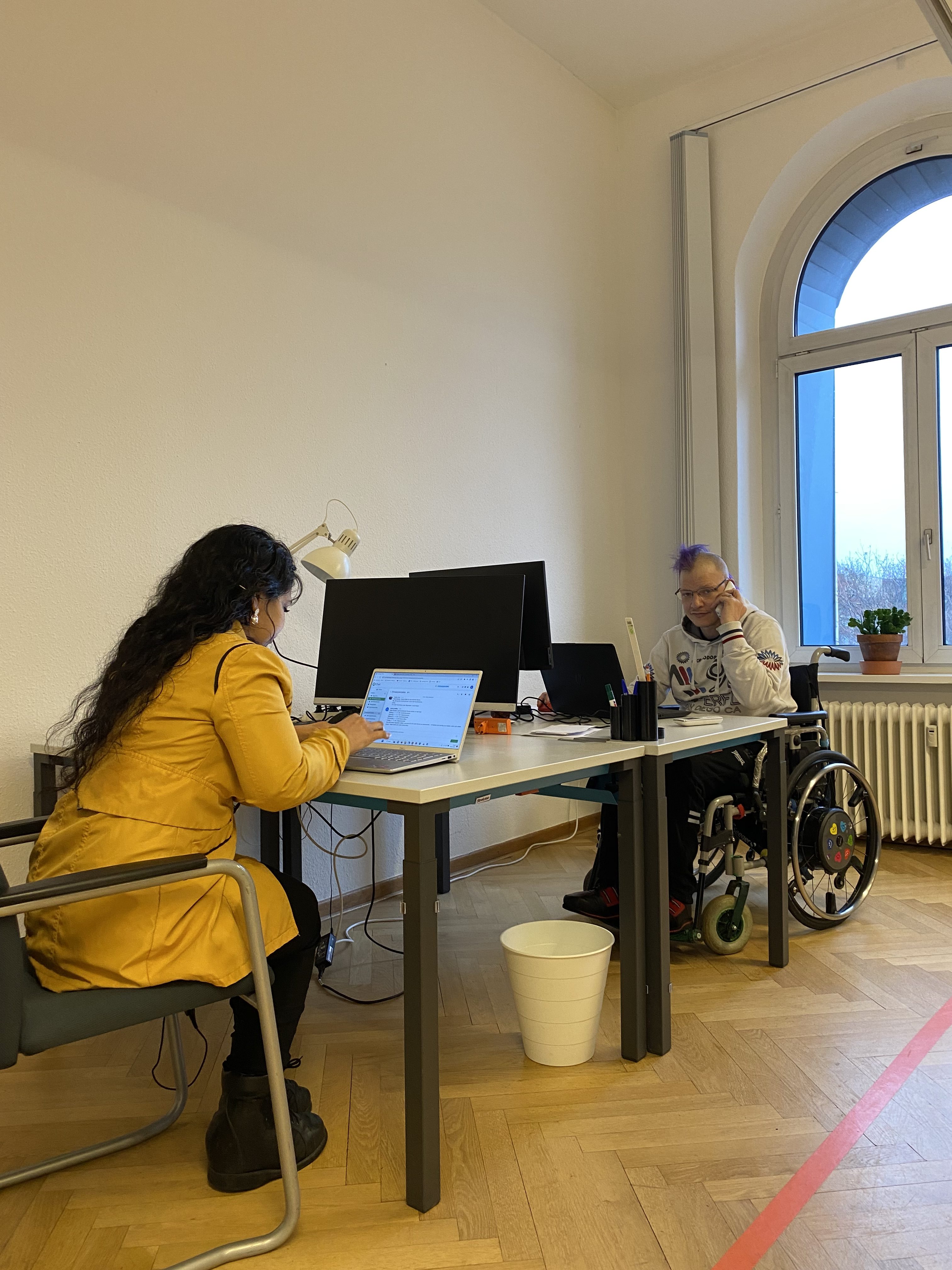 Im Bild sieht man einen freiwilligen Kollegen im Rollstuhl bei der telefonischen Kulturvermittlung und eine weitere Kollegin am Laptop. Beide sitzen an weißen Bürotischen in unserem barrierearmen Büro in Schöneberg.