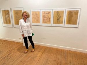 Die Künstlerin Jac Carley steht in einem Ausstellungsraum vor sechs abstrakten Bildern in den Farben Braun und Gold. Jac hat blonde gelockte Haare, sie trägt eine weiße Bluse und eine schwarze Hose.